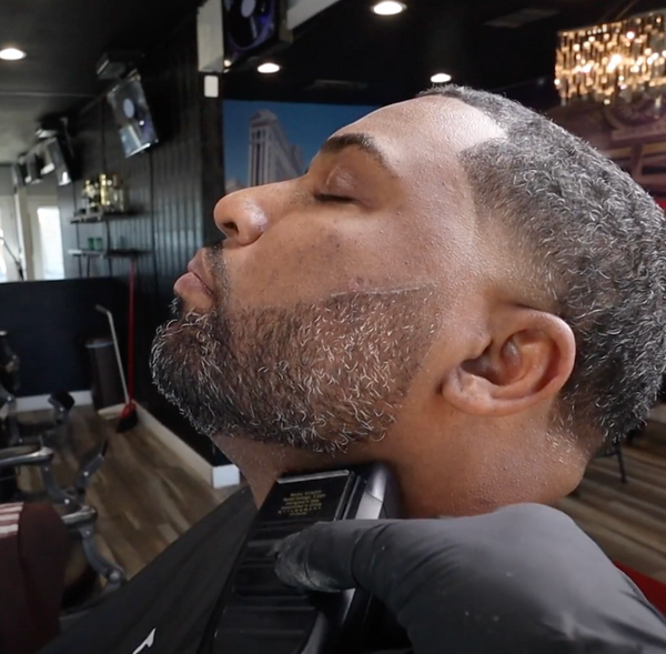 A close-up lifestyle shot of a man receiving a beard trim in a barbershop. His head is tilted back as a gloved hand uses an electric beard trimmer on his chin and jawline.