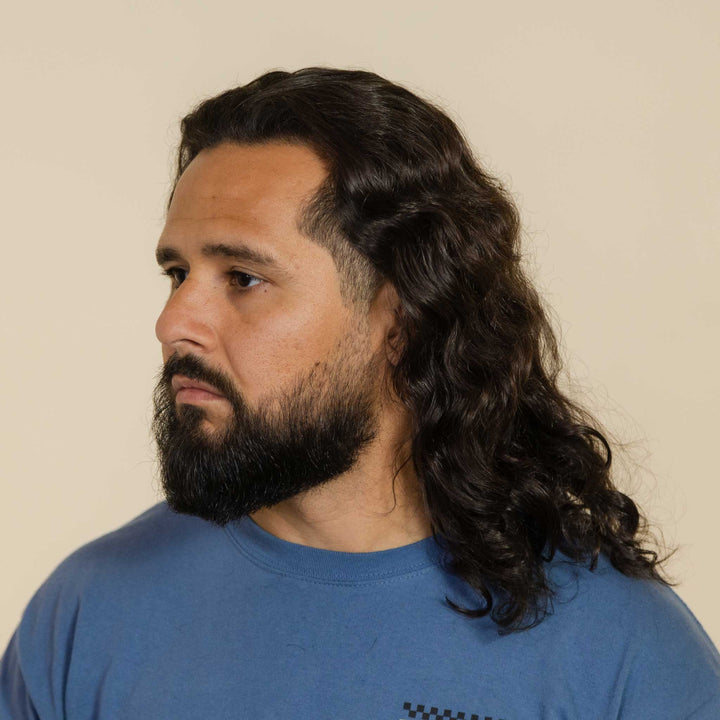 Man with long hair and beard wearing a blue shirt against a beige background