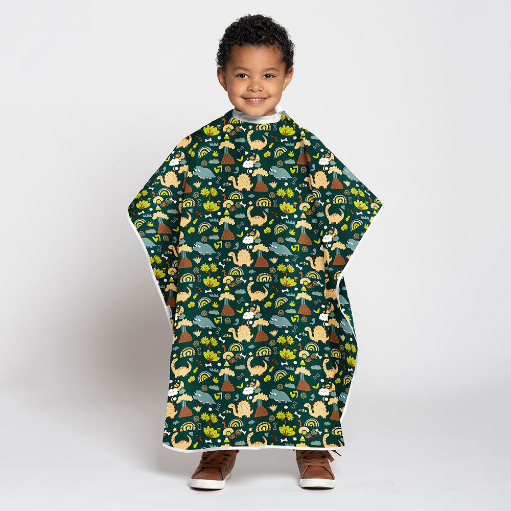 A young child wearing a dark green poncho-style blanket with a colorful cartoon dinosaur pattern, standing against a light gray studio background.