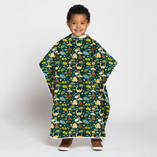 A young child wearing a dark green poncho-style blanket with a colorful cartoon dinosaur pattern, standing against a light gray studio background.