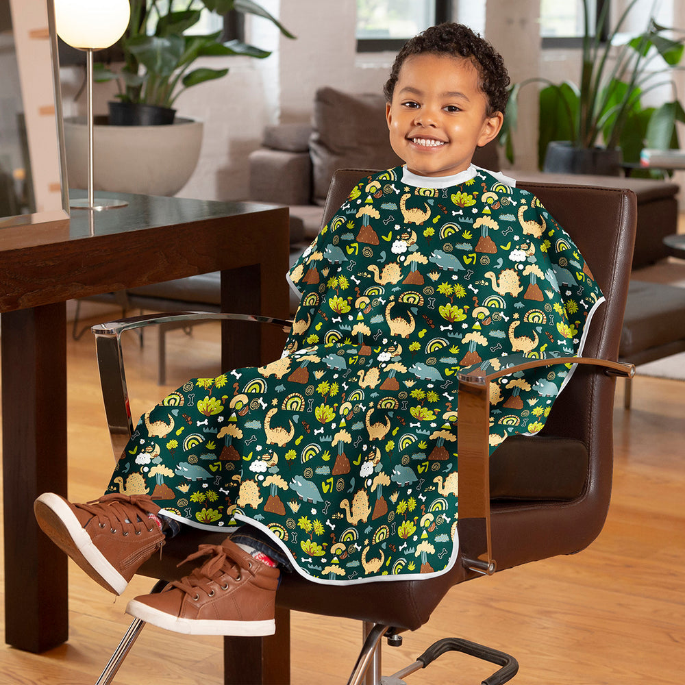 A young child wearing a green dinosaur-themed salon cape sits in a brown office chair in a living room setting.