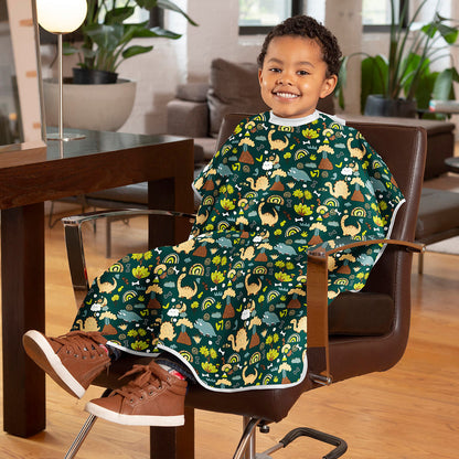A young child wearing a green dinosaur-themed salon cape sits in a brown office chair in a living room setting.