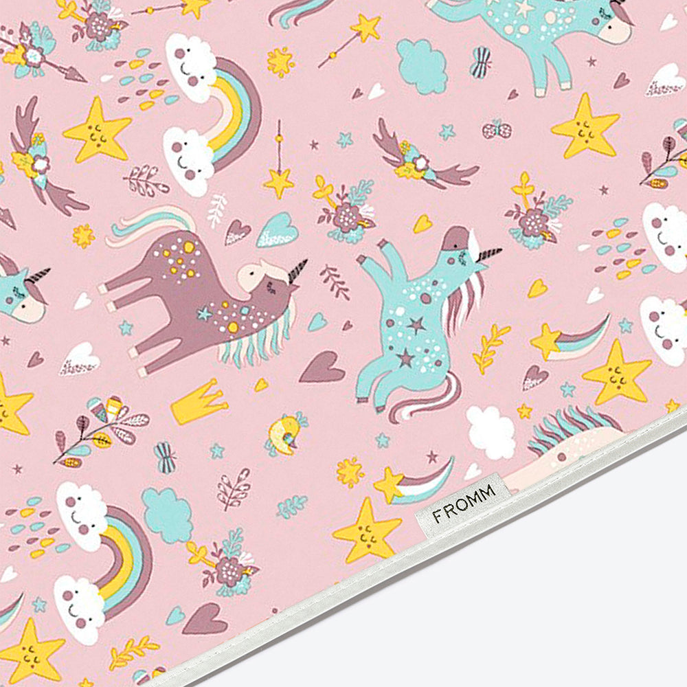 A close-up of a pink fabric cape printed with a whimsical unicorn pattern. The design features pastel pink unicorns, yellow stars, rainbows, clouds, and small decorative motifs.