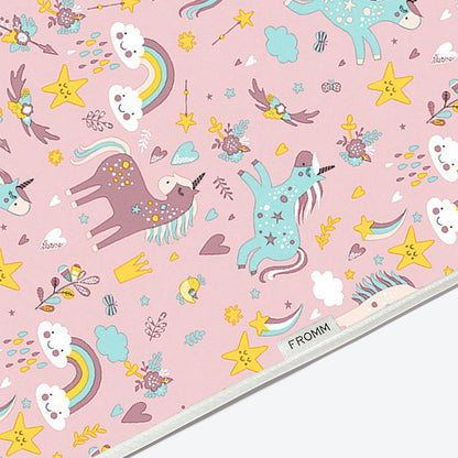 A close-up of a pink fabric cape printed with a whimsical unicorn pattern. The design features pastel pink unicorns, yellow stars, rainbows, clouds, and small decorative motifs.