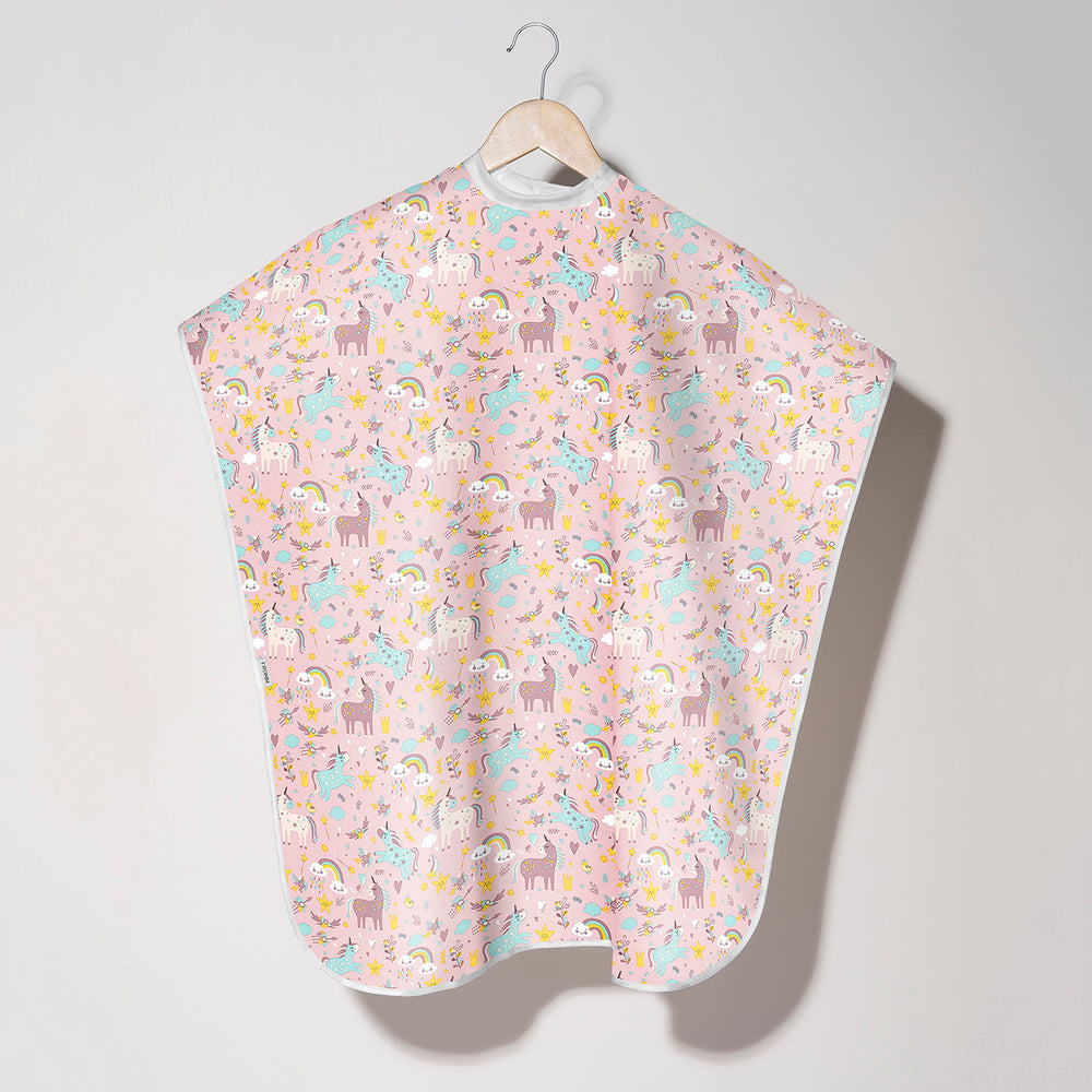 A kids cape with a light pink background and a playful pattern of cartoon unicorns, llamas, and rainbows, hanging on a wooden hanger against a neutral light gray wall.