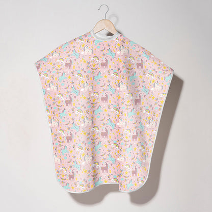 A kids cape with a light pink background and a playful pattern of cartoon unicorns, llamas, and rainbows, hanging on a wooden hanger against a neutral light gray wall.