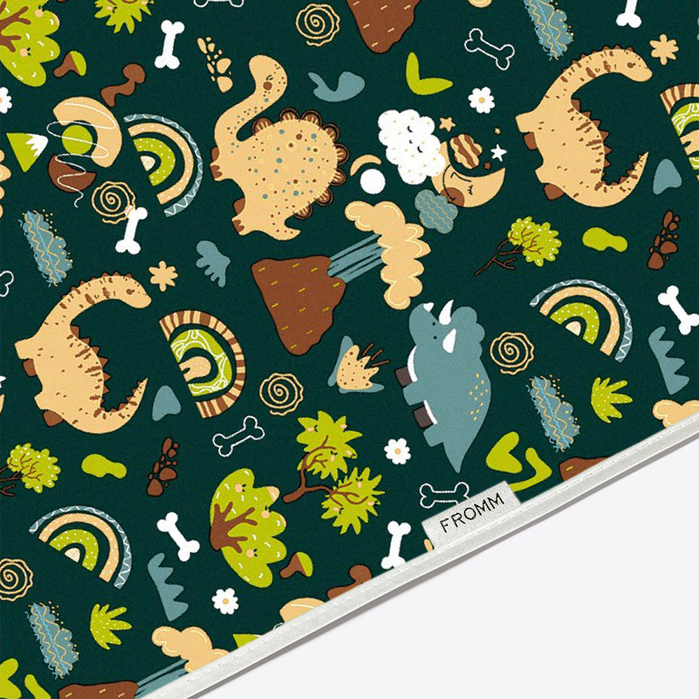 A close-up of a fabric or paper surface with a repeating whimsical dinosaur and nature print on a dark teal background. The pattern includes cartoon dinosaurs, trees, flowers, bones, and rainbows.