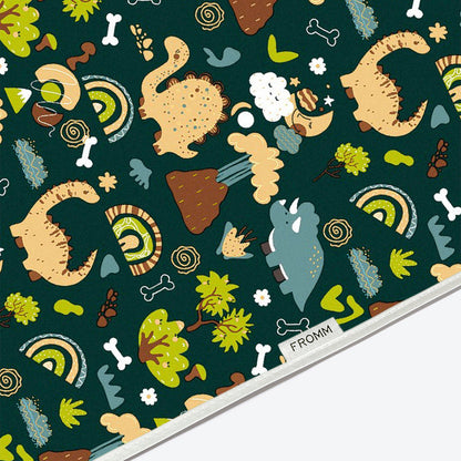 A close-up of a fabric or paper surface with a repeating whimsical dinosaur and nature print on a dark teal background. The pattern includes cartoon dinosaurs, trees, flowers, bones, and rainbows.