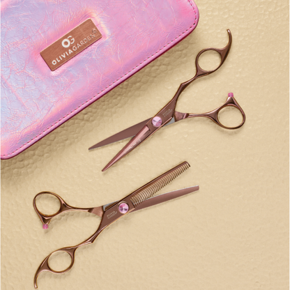two pink salon scissors with pink gem accents are arranged on a textured beige surface next to a pink iridescent pouch with a Olivia Garden logo.