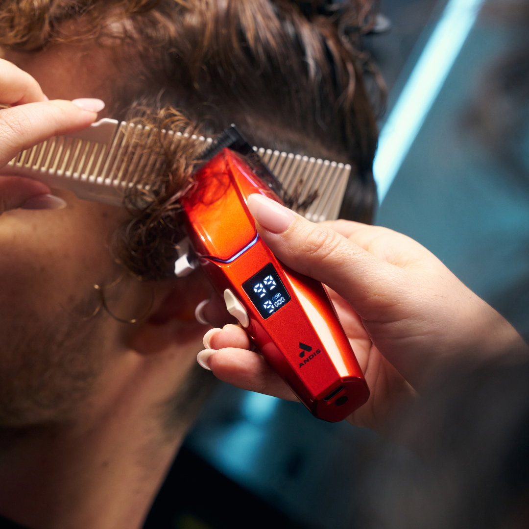 A close-up lifestyle shot of a person getting a haircut with a red and orange cordless electric clipper. A hand holds the clipper while another hand combs the hair.